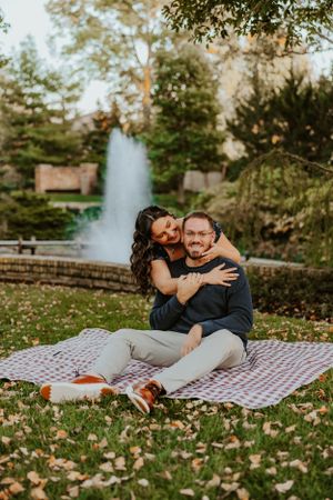 A couple enjoys a romantic autumn picnic on a blanket near a fountain in a scenic park setting.