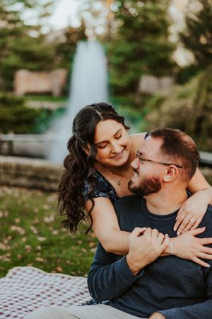 A couple shares an intimate moment while sitting on a picnic blanket in a park with trees in the background.