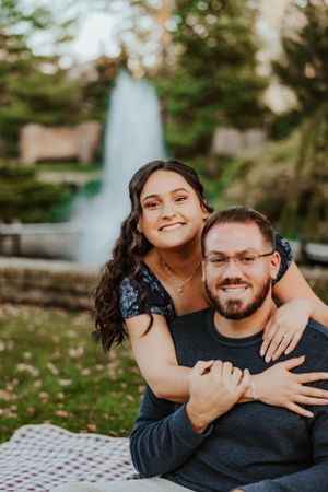 A happy couple embraces while sitting on a picnic blanket in a park with a fountain visible in the background.