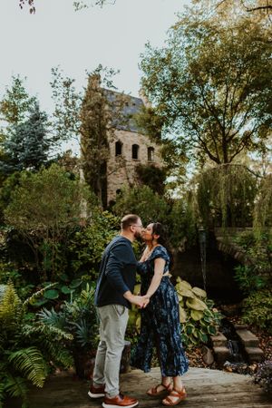 A couple shares a romantic kiss in a lush garden setting with an old stone tower in the background.