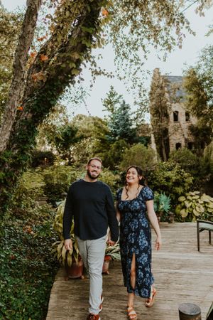 A couple walking and laughing together on a wooden pathway surrounded by lush greenery and trees near an old building.