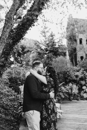 A romantic black and white photo of a couple embracing under a tree in a garden with a rustic building in background.