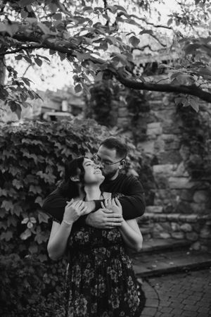 Romantic black and white photograph of a couple embracing under tree branches near a brick wall in a garden setting.