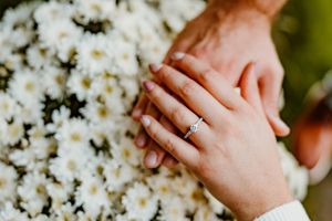 Hands wearing an engagement ring rest against a backdrop of white daisy flowers in soft focus.
