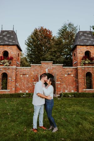 A romantic scene in front of a rustic brick wall with twin towers and autumn trees in the background.