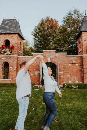 A couple dances together on grass in front of a rustic brick wall with two towers on an autumn day.