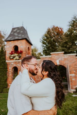 A romantic couple in white sweaters embrace in front of a rustic brick tower during autumn.
