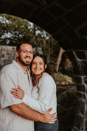 A couple embraces while standing under a stone archway during a warm sunset photoshoot.
