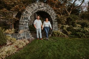 A couple holding hands stands in front of a stone archway surrounded by autumn foliage and green grass.