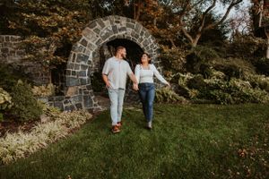 A couple walks together under a rustic stone archway surrounded by lush greenery during a golden hour photoshoot.