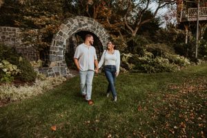 A couple stands holding hands in front of a rustic stone archway during golden hour in a garden setting.