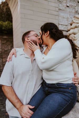 A romantic couple in casual attire share a tender intimate moment against a white brick wall backdrop.