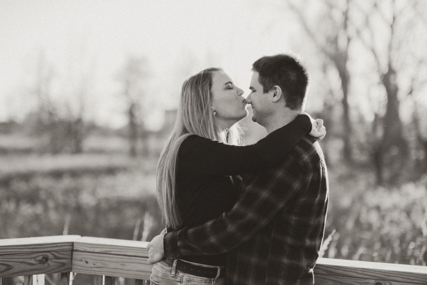 Black and white romantic outdoor portrait of a couple embracing and kissing while leaning against a wooden fence.