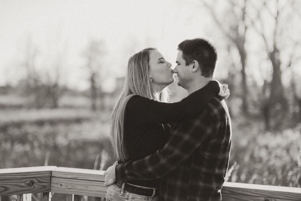 Black and white romantic outdoor portrait of a couple embracing and kissing while leaning against a wooden fence.