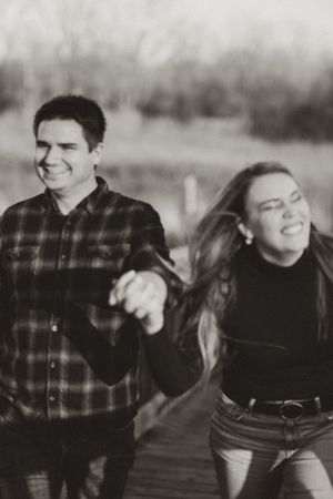 A joyful black and white photo of a couple running and laughing together on a wooden path outdoors.