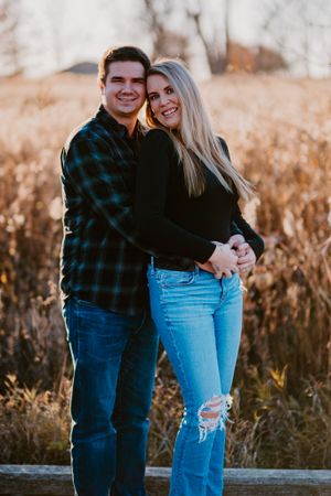 A couple embraces while standing in a field during golden hour, wearing casual attire of jeans and plaid.