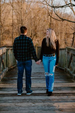 A couple holding hands walks across a wooden bridge in the autumn woods while wearing casual attire.