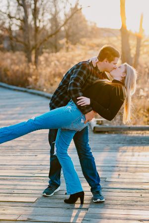 Romantic couple in casual attire sharing intimate moment on wooden boardwalk at sunset.