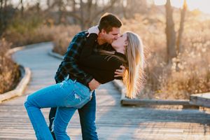 A romantic couple shares a kiss while posing on a wooden boardwalk at sunset.