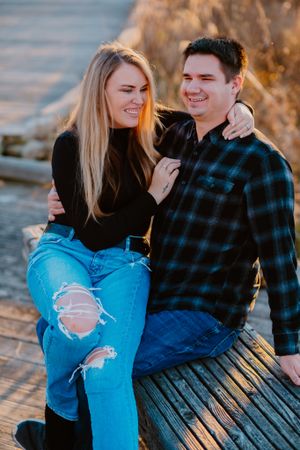 A couple laughs together while sitting on a wooden bench during golden hour, wearing casual denim and plaid.