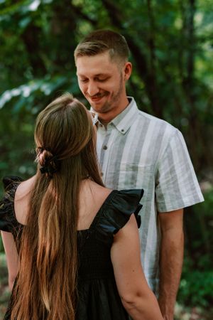 A romantic couple shares a tender moment in a wooded setting, with her long hair and his white shirt framed by lush greenery.