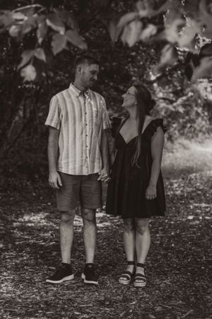 A couple in casual summer outfits stands together on a wooded path in black and white photography.