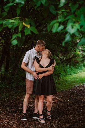A couple shares a romantic moment on a wooded path surrounded by lush green foliage.