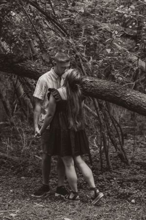 A couple embraces tenderly in a wooded area while leaning against a fallen tree in black and white photography.