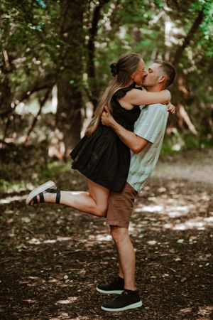 A romantic couple embraces and kisses on a wooded path as sunlight filters through the trees.