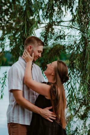 A romantic couple embraces under a drooping willow tree by the water on a peaceful summer evening.