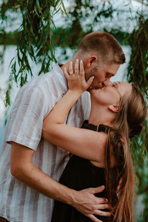 A romantic couple shares an intimate kiss under hanging willow branches by a lake.