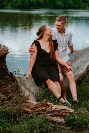 A couple sits together on a fallen log by a serene lake, sharing a joyful moment in nature.