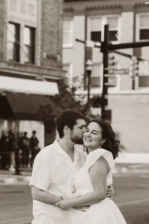 A romantic black and white street scene shows a tender embrace between two people dressed in white on a city corner.