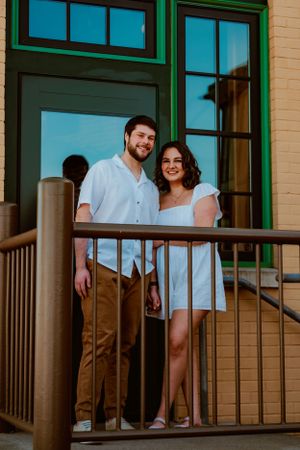 A smiling couple stands on a wooden deck by green-trimmed windows of a beige building in matching white outfits.