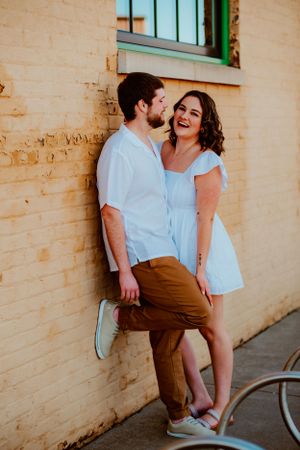 Couple in light blue and brown outfits sharing a candid laughing moment against a beige wall during a photoshoot.