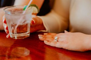 A glass of cocktail with ice and lime slice sits on a wooden table next to a hand wearing a silver ring.