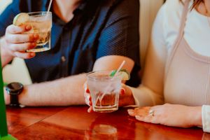 Two people sitting at a wooden table holding glasses of whiskey or bourbon with ice cubes.