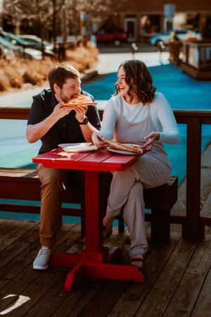 Two people sharing pizza and laughing while sitting at a red picnic table on a wooden deck during golden hour.
