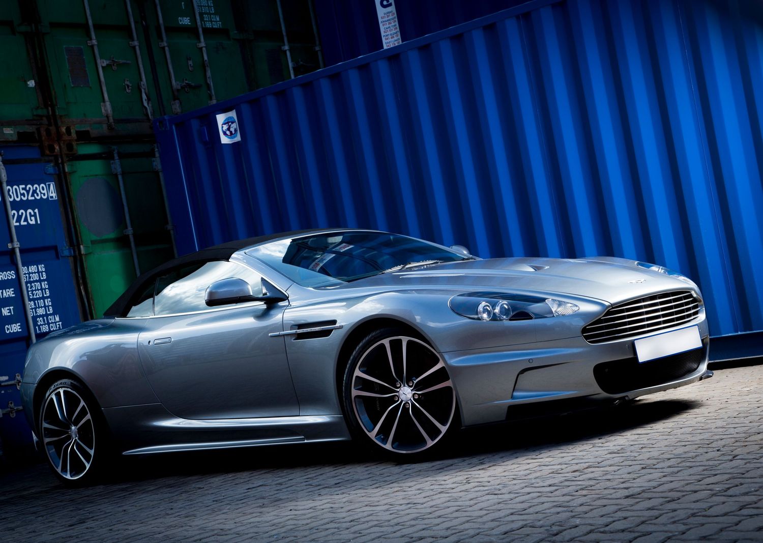 A sleek silver Aston Martin DBS sports car parked in front of blue shipping containers.