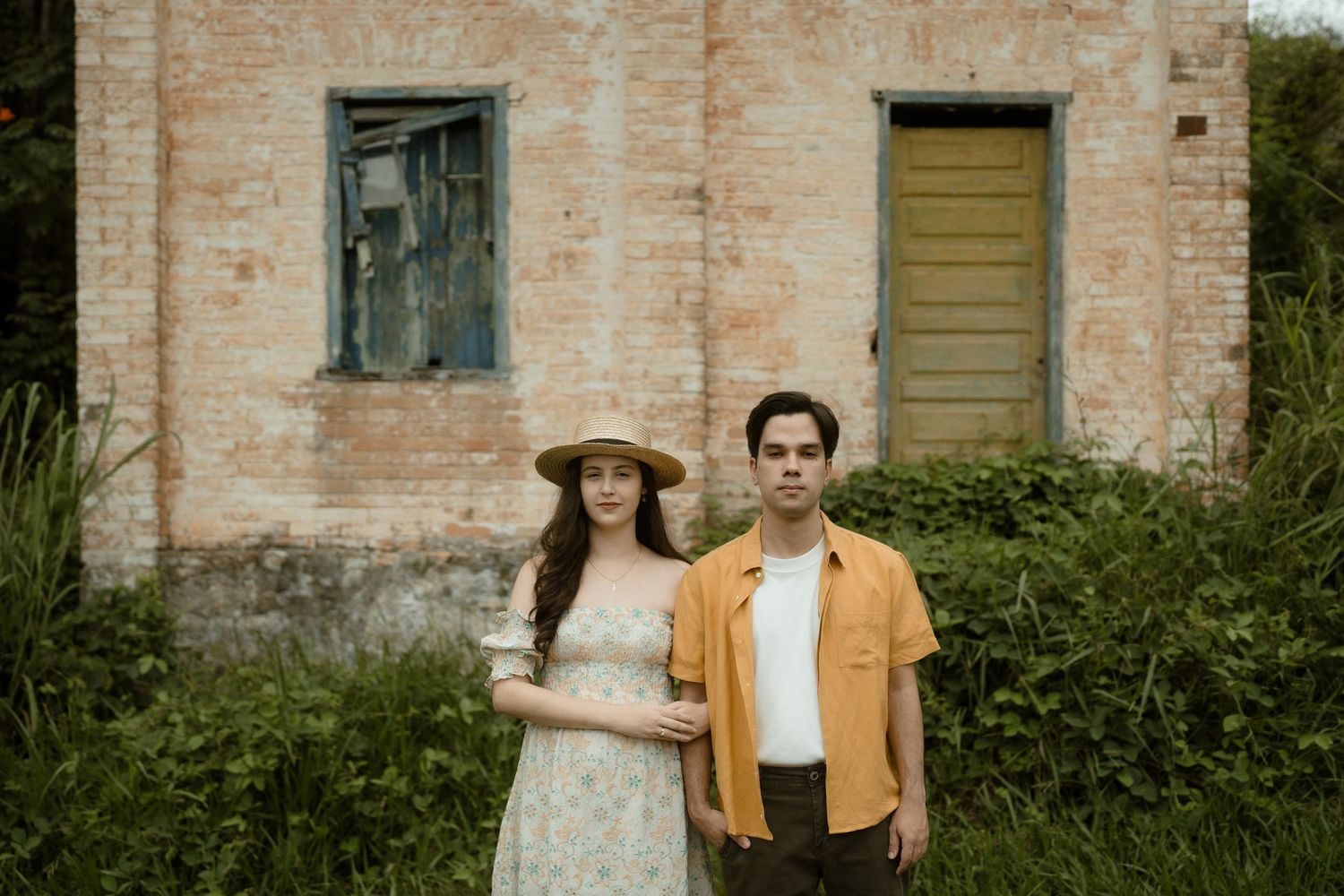 A couple poses against a rustic brick building with vintage doors and windows.