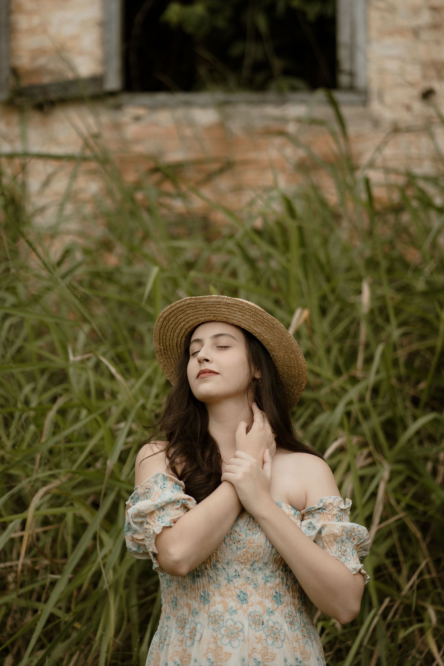Woman in floral dress and straw hat poses expressively by tall grass.
