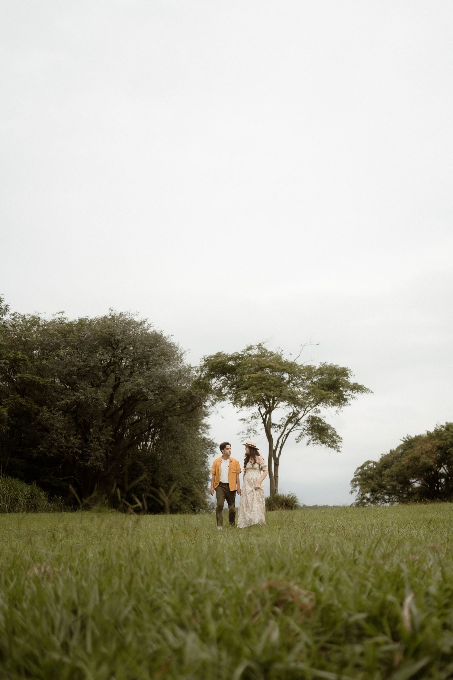 A couple stands together in a grassy field with trees silhouetted against an overcast sky.