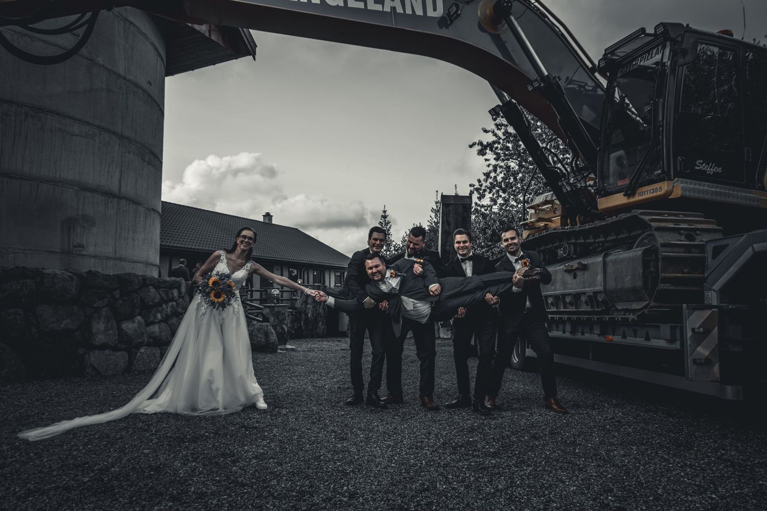 Wedding party poses playfully in front of large industrial crane at unique outdoor venue.
