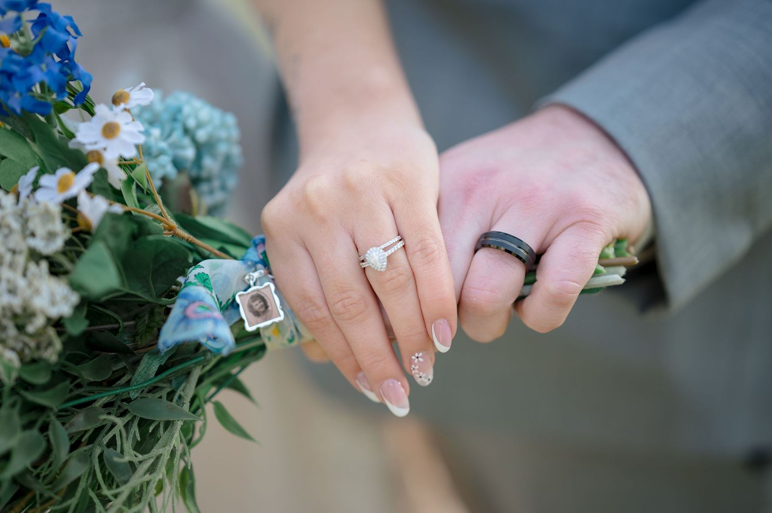 Two hands with wedding rings hold a blue and white floral bouquet against a soft background.