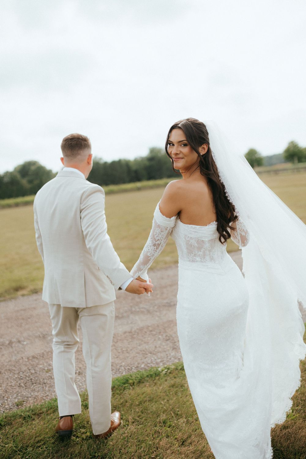 Bride in white lace gown and veil looks back smiling while holding groom's hand outdoors.