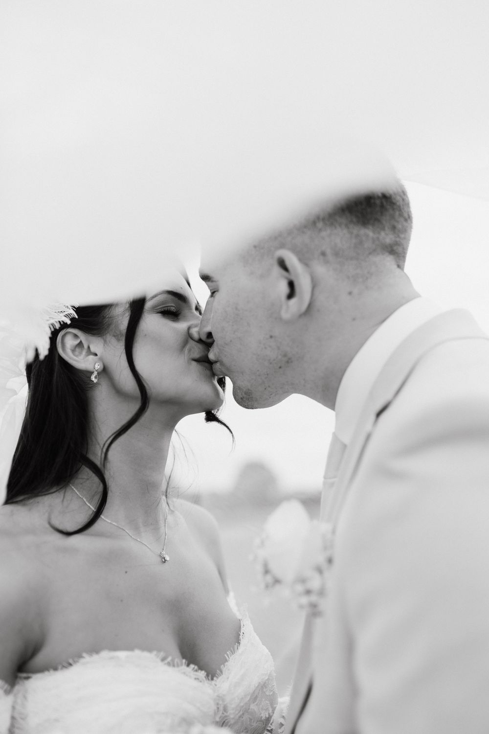 Black and white photo of a bride and groom sharing a romantic kiss under a veil on their wedding day.