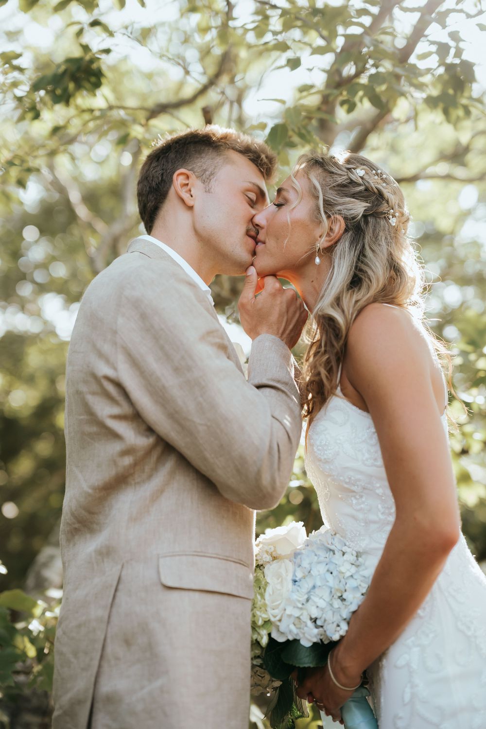 Bride and groom sharing a kiss outdoors under trees, groom in tan suit, bride holding white hydrangea bouquet.