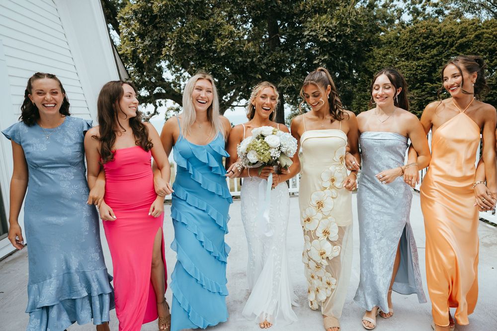 Bride in white floral gown walks with six bridesmaids in colorful mismatched dresses outdoors.