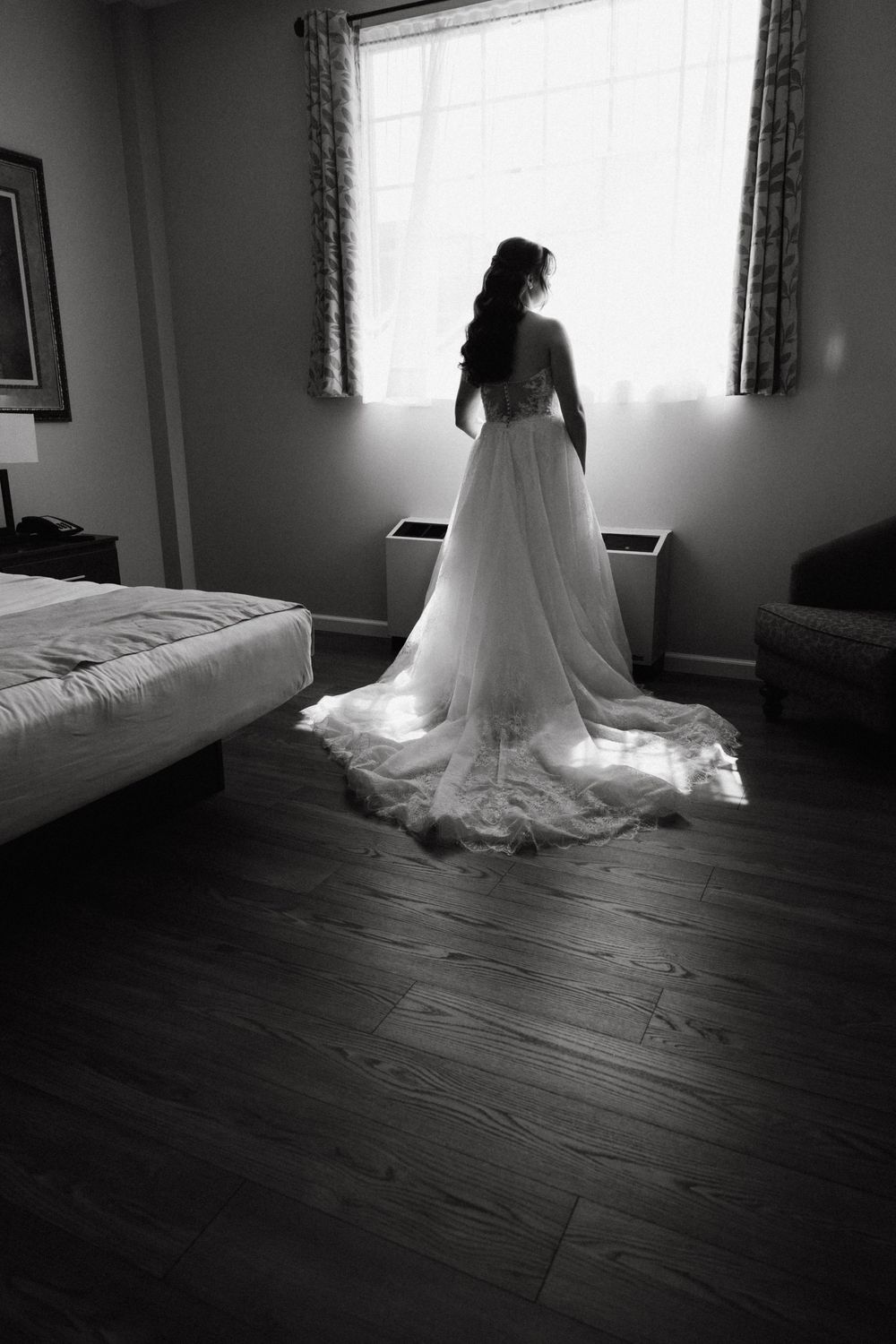 Black and white photo of a bride in a flowing wedding gown with a long train standing before a sunlit window.