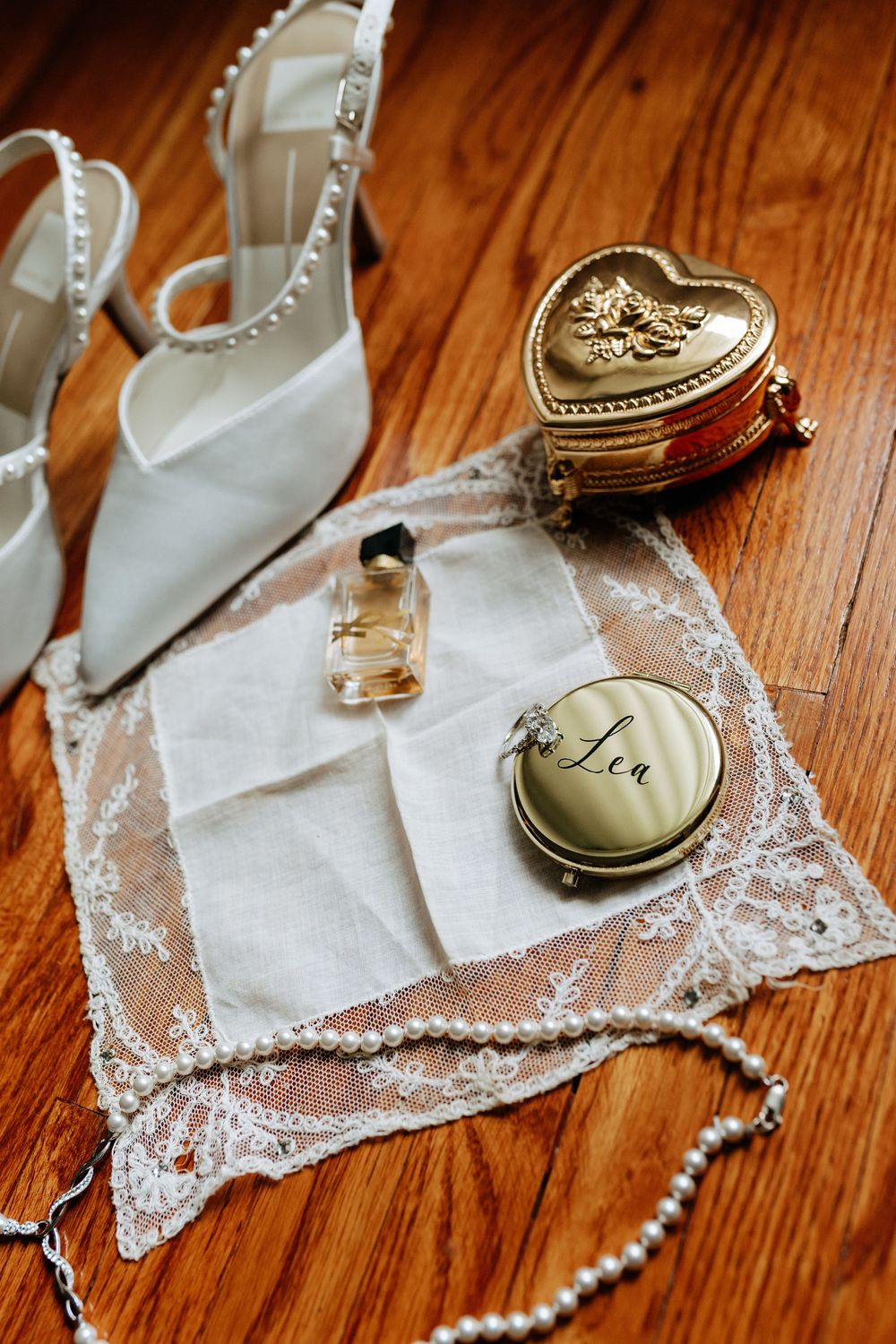 Elegant bridal accessories flat lay with pearl heels, heart jewelry box, perfume, compact mirror, lace handkerchief, and pearl necklace.